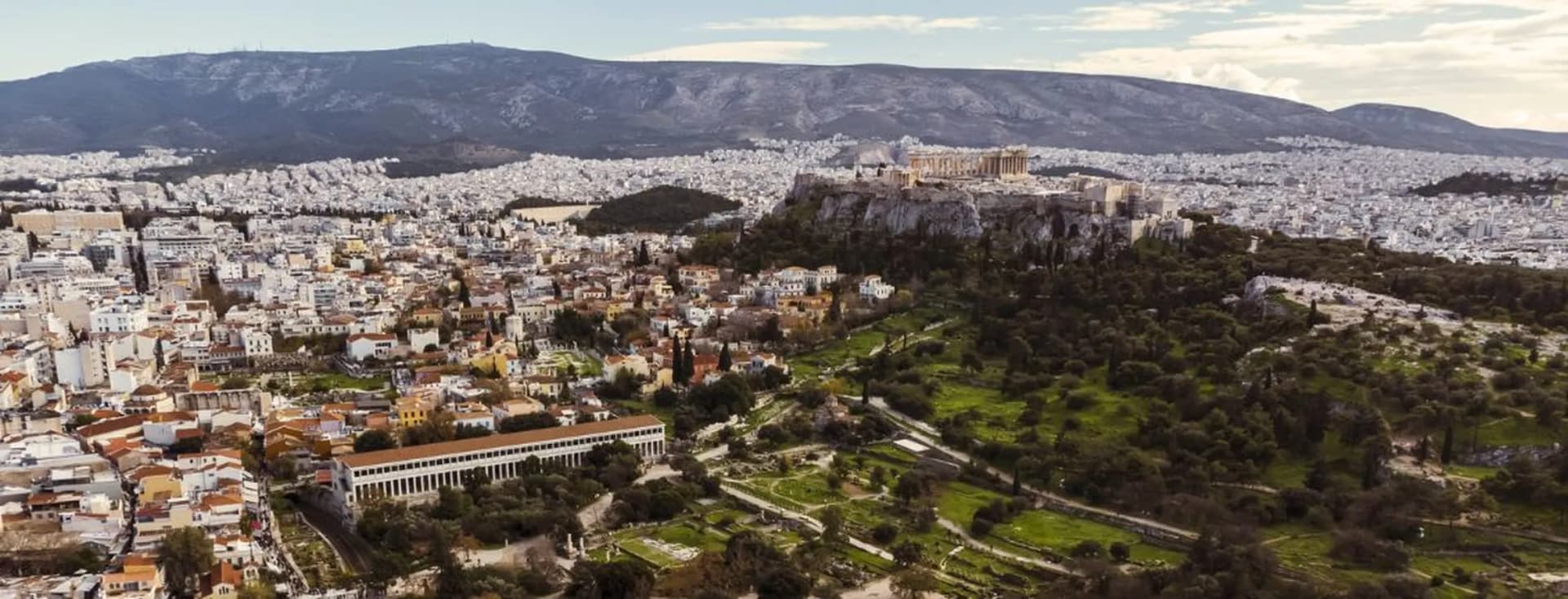 Aerial view of Athens with the Parthenon atop the Acropolis, sprawling city streets, green hills in the foreground, and distant mountains.
