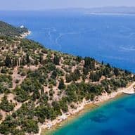 Aerial view of a large, wooded peninsula jutting into deep blue waters, featuring a white villa nestled among the trees and a small island just offshore.