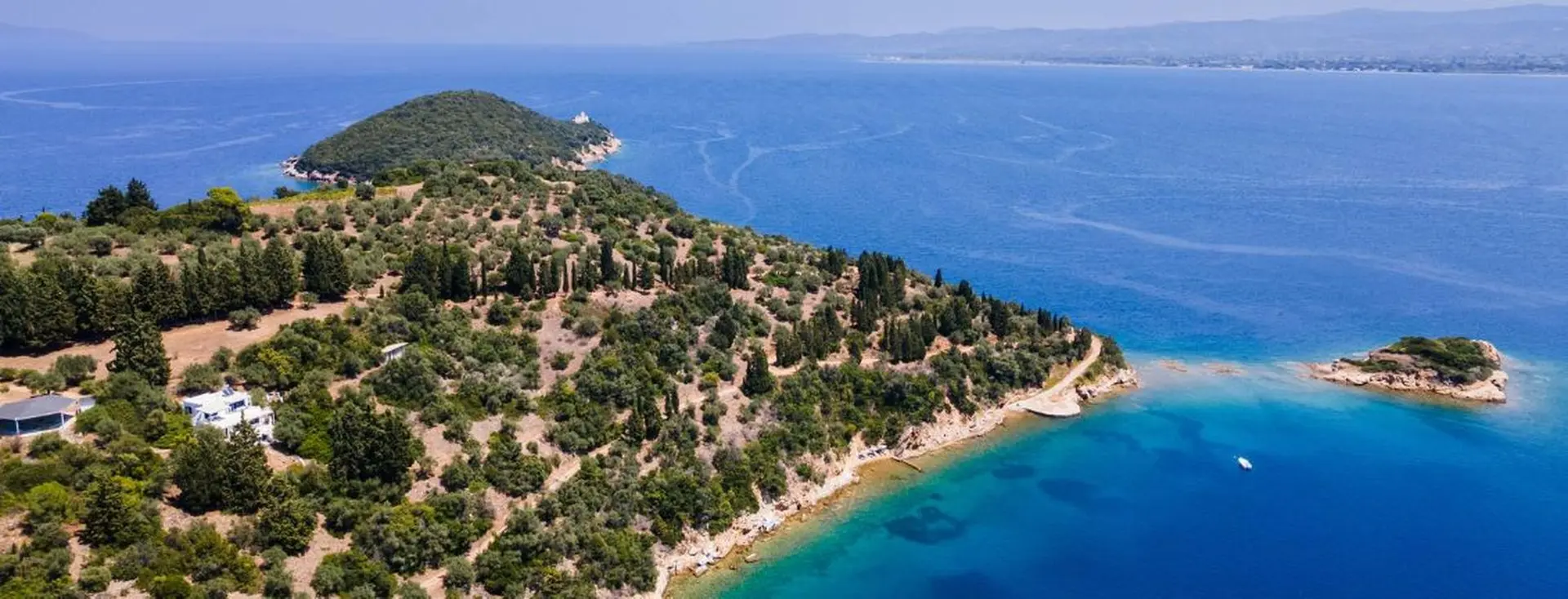 Aerial view of a large, wooded peninsula jutting into deep blue waters, featuring a white villa nestled among the trees and a small island just offshore.