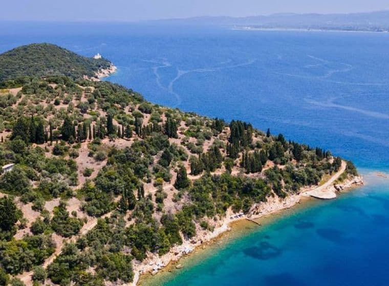 Aerial view of a large, wooded peninsula jutting into deep blue waters, featuring a white villa nestled among the trees and a small island just offshore.