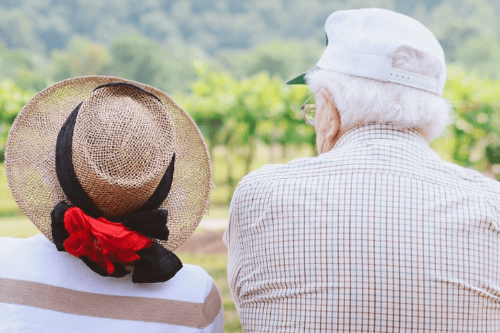 Elderly couple sitting on a bench, wearing hats, facing a scenic view with greenery and trees in the background