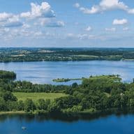 Luftaufnahme von blauen Seen und Inseln, umrahmt von grünen Wäldern, Feldern und einem kleinen Dorf unter einem teilweise bewölkten Himmel