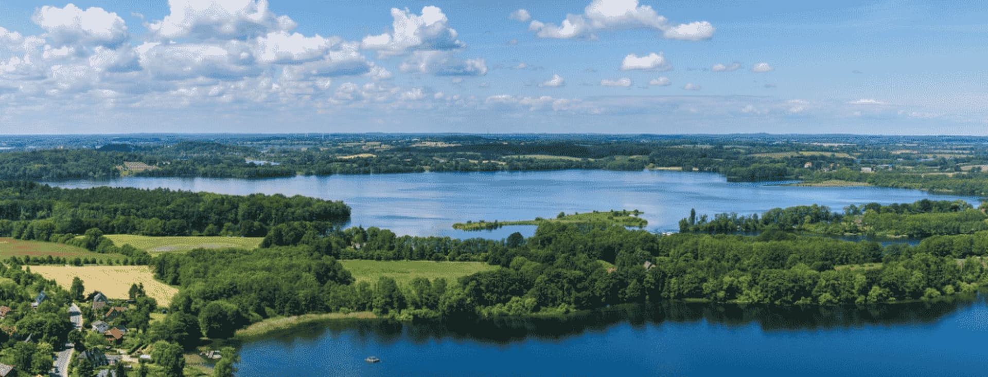 Luftaufnahme von blauen Seen und Inseln, umrahmt von grünen Wäldern, Feldern und einem kleinen Dorf unter einem teilweise bewölkten Himmel
