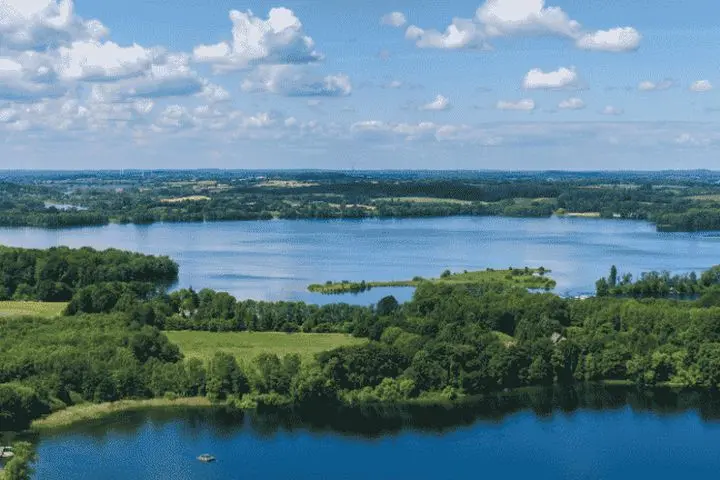 Luftaufnahme von blauen Seen und Inseln, umrahmt von grünen Wäldern, Feldern und einem kleinen Dorf unter einem teilweise bewölkten Himmel