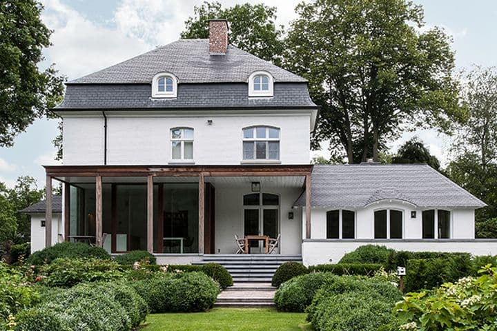 Elegant white house with modern additions and landscaped garden, showcasing how aesthetic improvements contribute to increasing property value. The home features a slate roof, arched windows, and a glass-enclosed porch, surrounded by lush greenery and manicured shrubs, illustrating effective ways to boost a property's market appeal and worth.