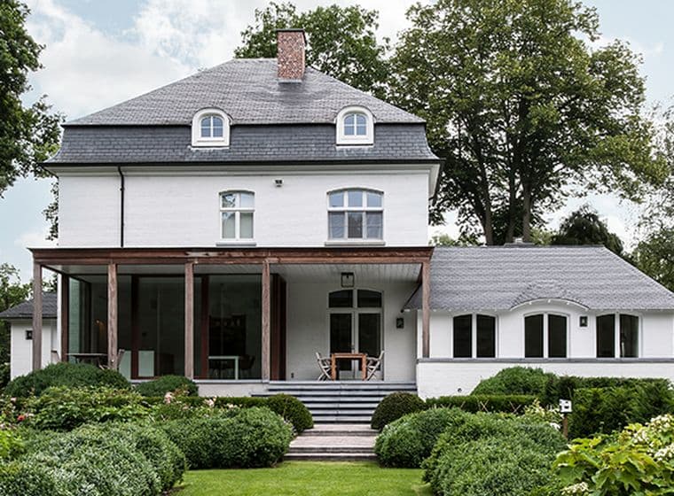 Zweistöckiges weißes Haus mit grauem Dach, umgeben von grünen Bäumen und Büschen. Eine verglaste Veranda ist sichtbar.