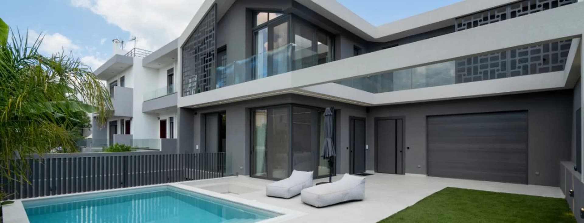 Modern two-story villa exterior with dark grey facade, swimming pool, outdoor sun loungers, glass railing balconies, and garage door