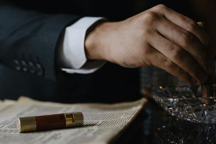 A person in a suit holding a cigar over an ashtray, with an open book and a cigar case nearby.
