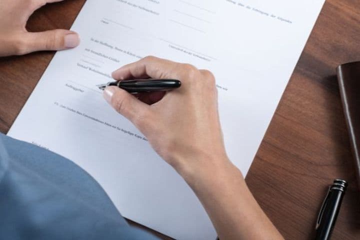 A person signing a document on a wooden desk with a black pen, next to a leather wallet and a set of keys.