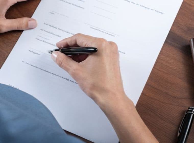 A person signing a document on a wooden desk with a black pen, next to a leather wallet and a set of keys.