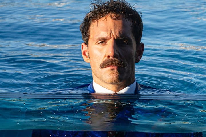 Άντρας με μουστάκι, φορώντας κοστούμι, στέκεται μέσα στο νερό μέχρι το ύψος του στήθους, με φόντο μια ήρεμη γαλάζια θάλασσα.