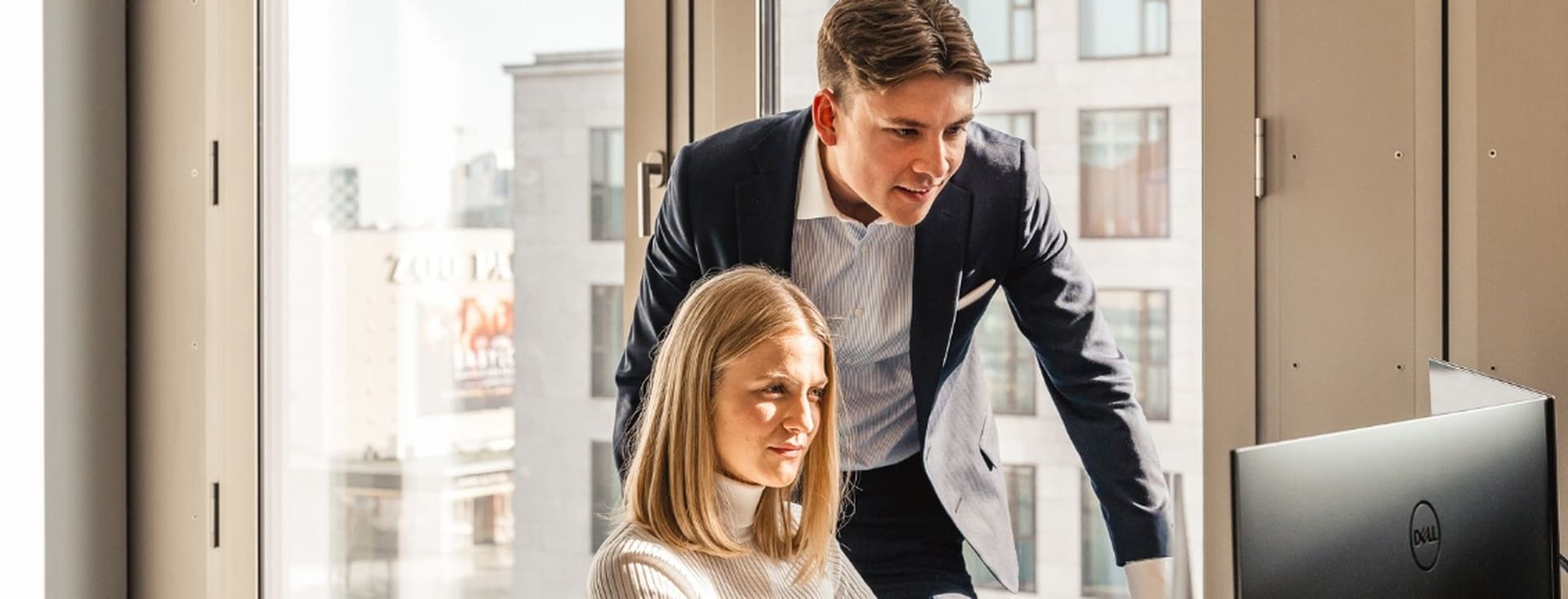 A man in a suit leans over a computer screen, discussing with a woman seated at a desk in a modern office.
