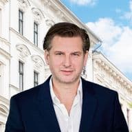 Man in navy blazer and white shirt stands before an ornate white apartment building with blue sky and a leafy tree.