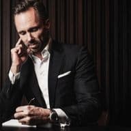 Man in a suit sits at a table, talking on the phone and writing in a notebook. Dark, elegant setting with vertical paneling in the background.