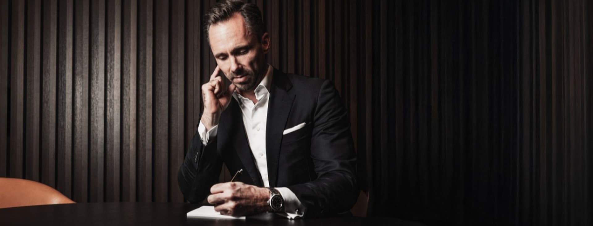 Man in a suit sits at a table, talking on the phone and writing in a notebook. Dark, elegant setting with vertical paneling in the background.