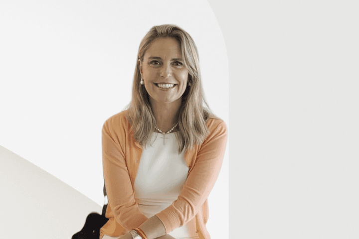 A smiling woman with blonde hair wearing a white dress and orange cardigan.