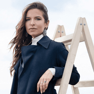 Γυναίκα με μαύρο κομψό παλτό ποζάρει δίπλα σε ξύλινη σκάλα σε μινιμαλιστικό θαλάσσιο σκηνικό.