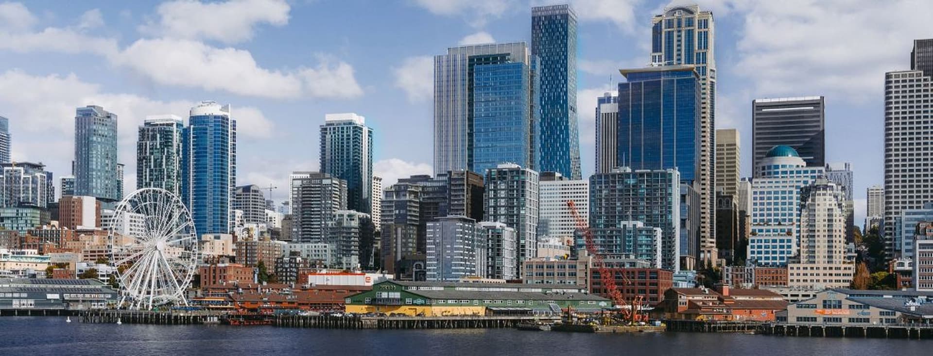 City skyline along the waterfront, featuring a large white Ferris wheel and tall glass skyscrapers against a blue sky.