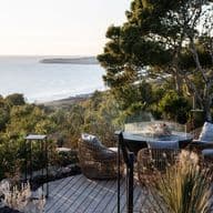 Wooden seaside deck with woven lounge chairs and a round fire table, overlooking coastal shrubs, trees and a sunlit beach at sunset.