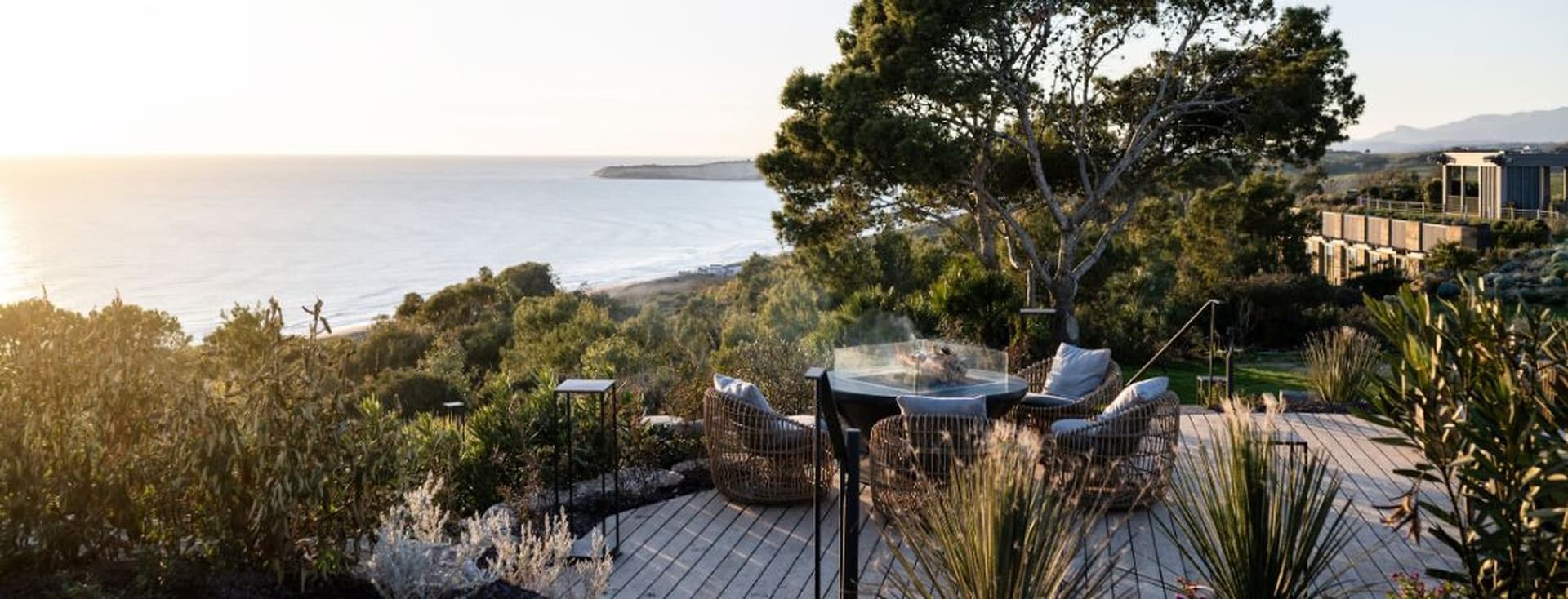 Eine Holzterrasse mit Liegestühlen aus Korbgeflecht rund um eine Feuerstelle aus Glas, mit Blick auf die Küstenvegetation und das Meer bei Sonnenuntergang.