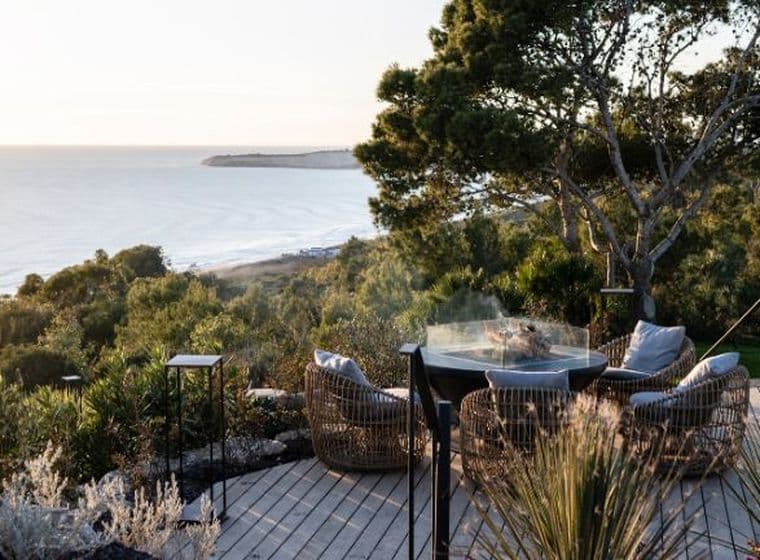 Wooden seaside deck with woven lounge chairs and a round fire table, overlooking coastal shrubs, trees and a sunlit beach at sunset.