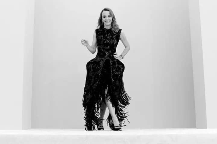 Woman in a black feathered gown poses center against a white backdrop; hands on hips, Netflix logos visible.