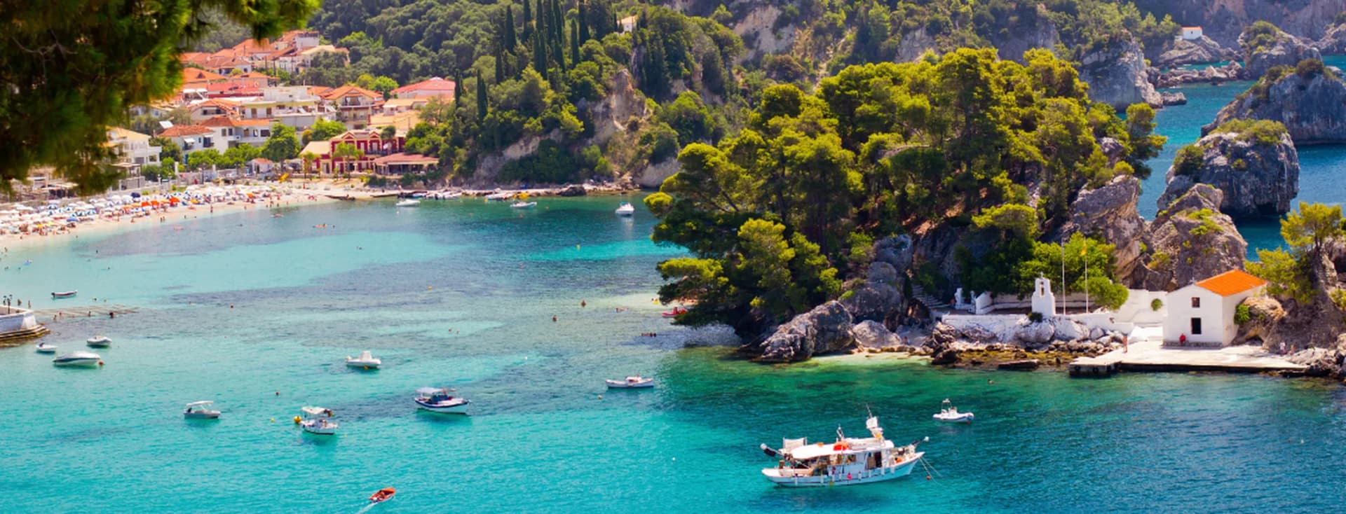 Scenic coastal bay in Parga with turquoise waters, boats, sandy beach, pine-covered islet, and white church.