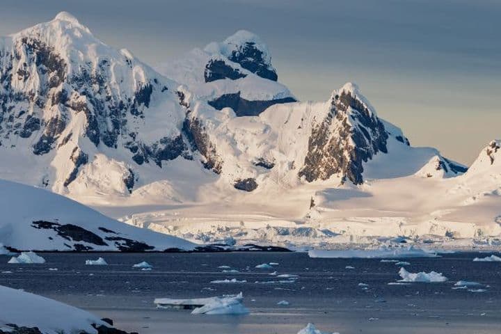 Snow-covered mountains and glaciers by a calm sea, with floating icebergs under a cloudy sky in a polar landscape.