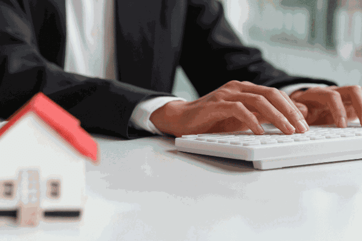 Person in a suit typing on a keyboard at a desk, with a small model house in the foreground.