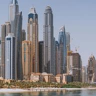 Skyline of a Dubai Marina housing market with tall skyscrapers, a waterfront, a beach, and clear blue skies.
