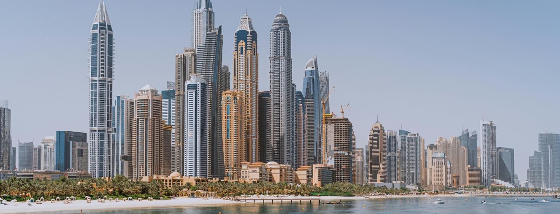 Skyline of a Dubai Marina housing market with tall skyscrapers, a waterfront, a beach, and clear blue skies.