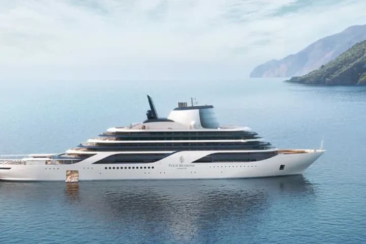 A white luxury yacht on the calm blue sea, with the green, mountainous coastline in the background.