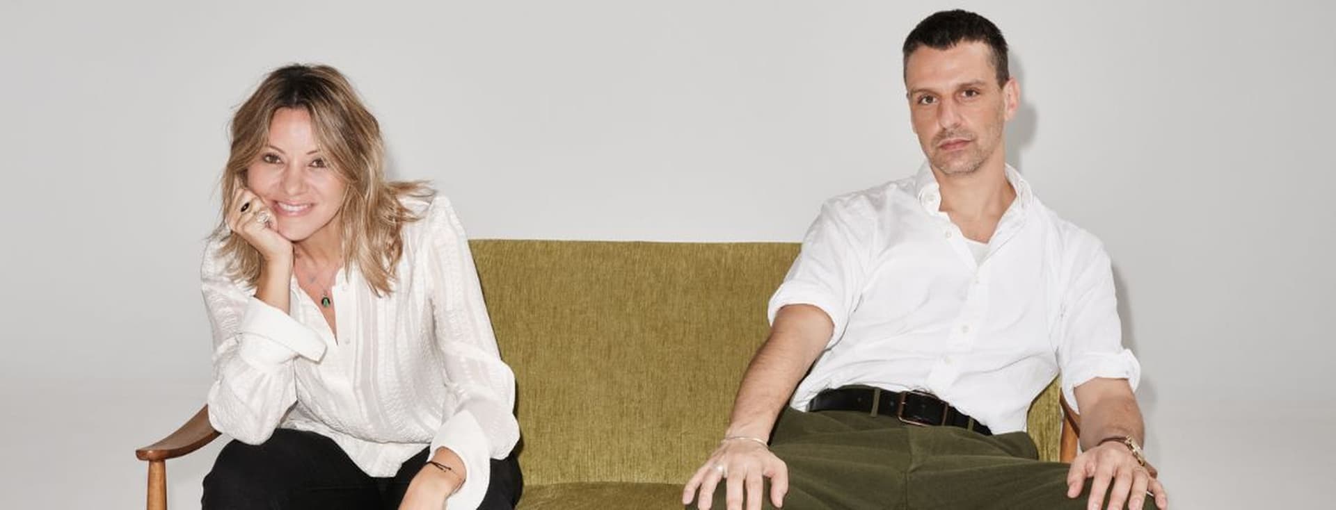 Two people are sitting on a green sofa against a plain background. Both are dressed casually in white tops and are looking at the camera.