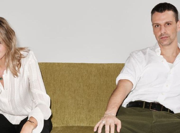 Zwei Personen sitzen auf einem grünen Sofa vor einem schlichten Hintergrund. Beide sind leger in weißen Oberteilen gekleidet und blicken in die Kamera.