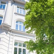 Low-angle view of a white historic building with arched windows and decorative facade, framed by a leafy green tree and bright blue sky.
