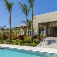 Modern luxury villa with a swimming pool, sun loungers, palm trees, and lush tropical landscaping under a clear blue sky.