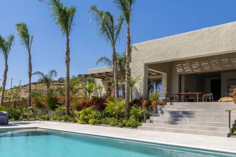 Modern luxury villa with a swimming pool, sun loungers, palm trees, and lush tropical landscaping under a clear blue sky.
