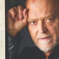 Portrait of a middle-aged man with a thoughtful expression, leaning against a bookshelf next to a cookbook, in a softly lit interior.