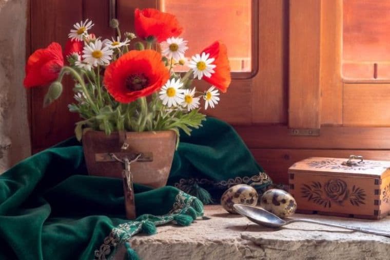 Rustic windowsill still life with red poppies, daisies, green cloth, and wooden music box in warm sunlight.