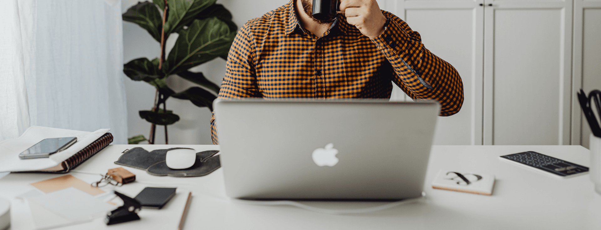 A person in a plaid shirt drinking from a mug at a white desk with a laptop and office supplies.