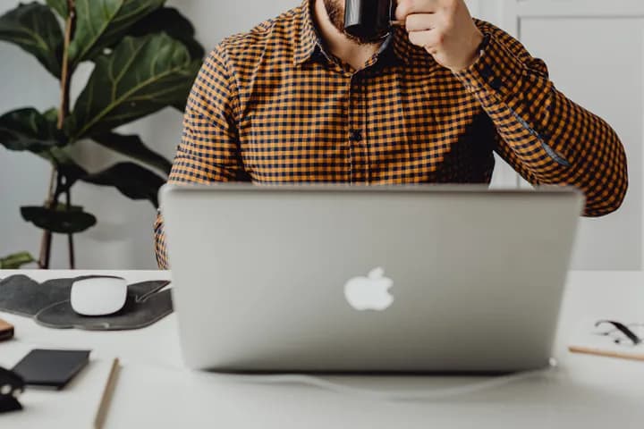A person in a plaid shirt drinking from a mug at a white desk with a laptop and office supplies.