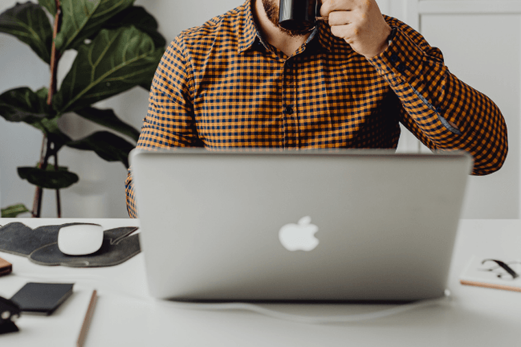 A person in a plaid shirt drinking from a mug at a white desk with a laptop and office supplies.