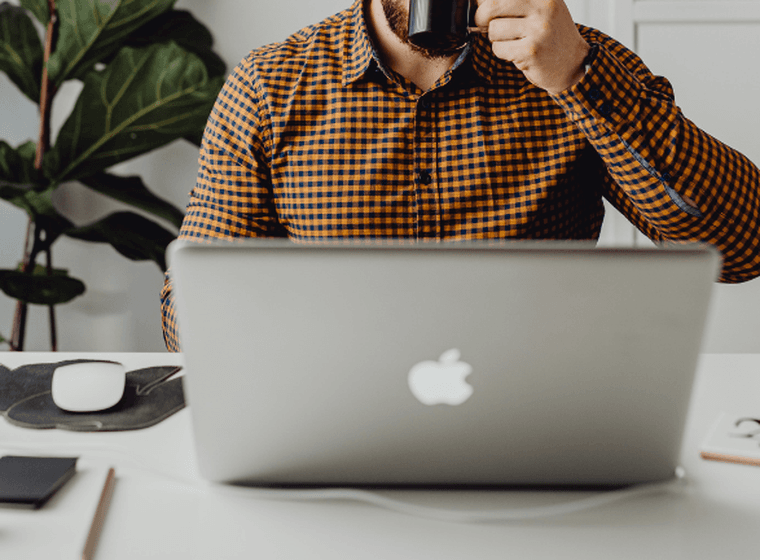 A person in a plaid shirt drinking from a mug at a white desk with a laptop and office supplies.