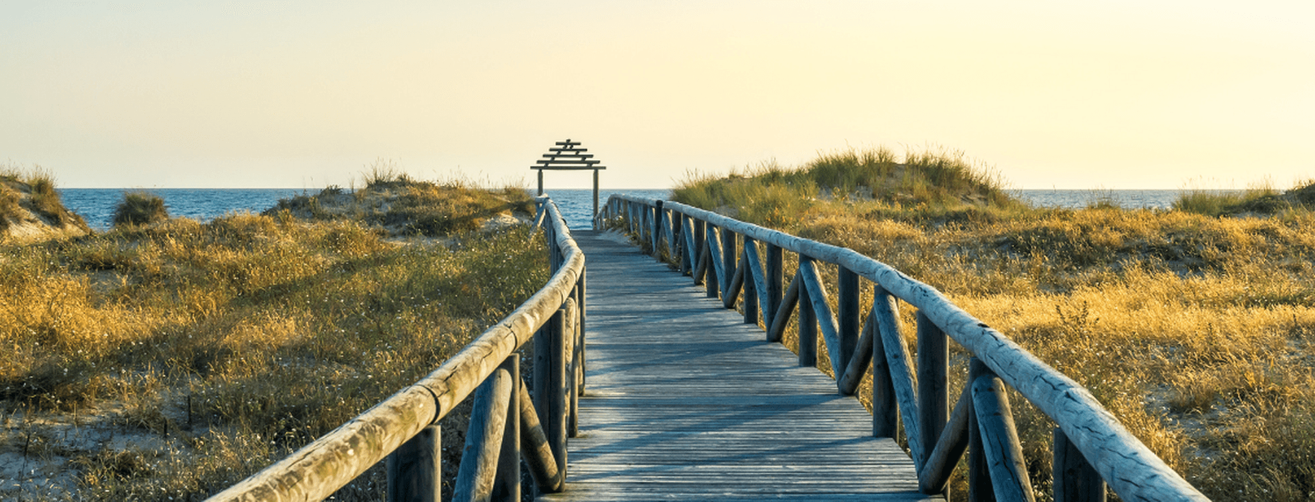 Un pontó de fusta és l'entrada a la platja, amb vegetació salvatge a banda i banda.
