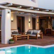 An outdoor Mediterranean-style terrace with a wooden pergola, white columns, wooden lounge furniture, terracotta floor tiles, and a swimming pool.