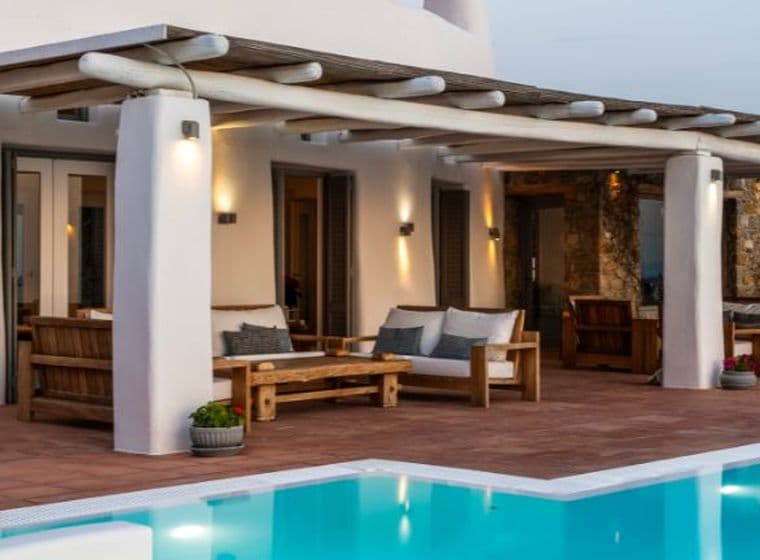 An outdoor Mediterranean-style terrace with a wooden pergola, white columns, wooden lounge furniture, terracotta floor tiles, and a swimming pool.