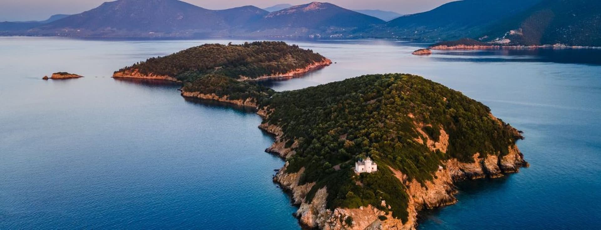 Aerial view of a long, heavily wooded peninsula with a lighthouse at the point, set against distant mountains.