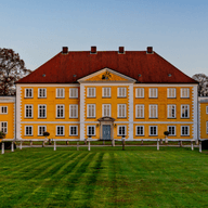 Ein gelbes, historisches Anwesen mit rotem Ziegeldach, einem zentralen Giebel und zwei Flügeln, das hinter einem gepflegten grünen Rasen und kahlen Bäumen liegt