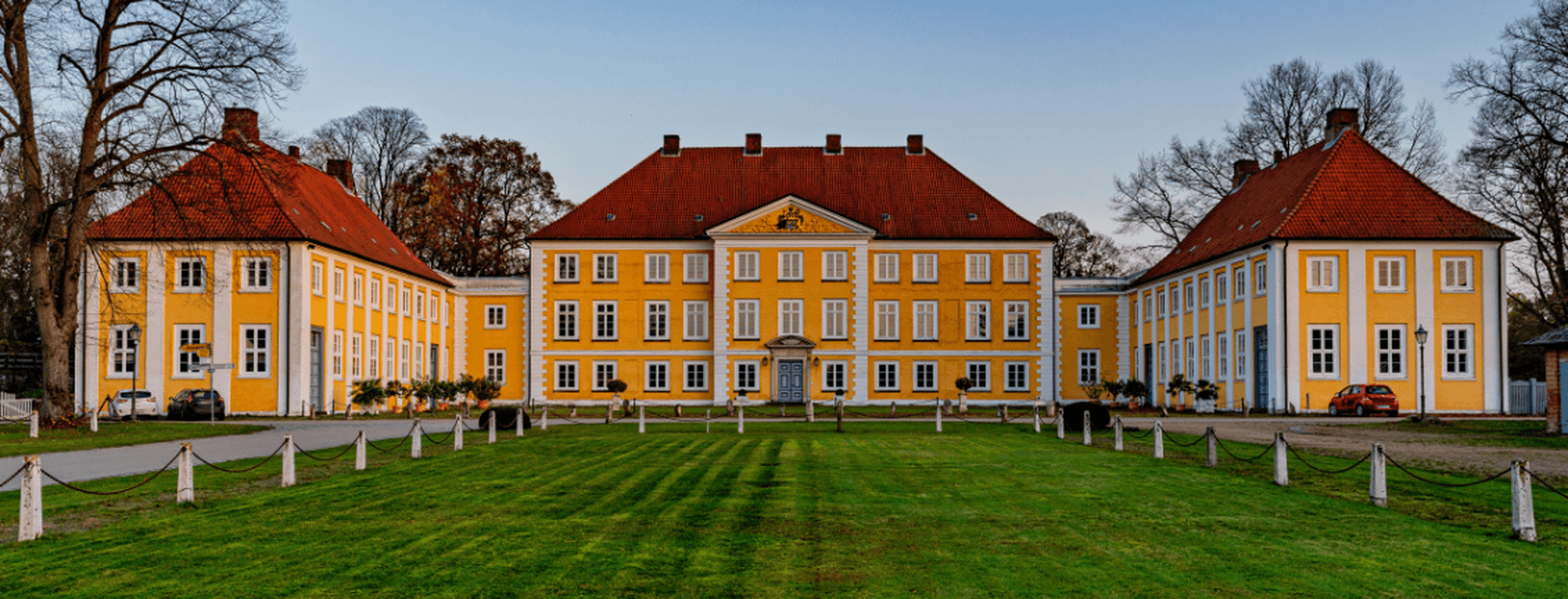 Ein gelbes, historisches Anwesen mit rotem Ziegeldach, einem zentralen Giebel und zwei Flügeln, das hinter einem gepflegten grünen Rasen und kahlen Bäumen liegt