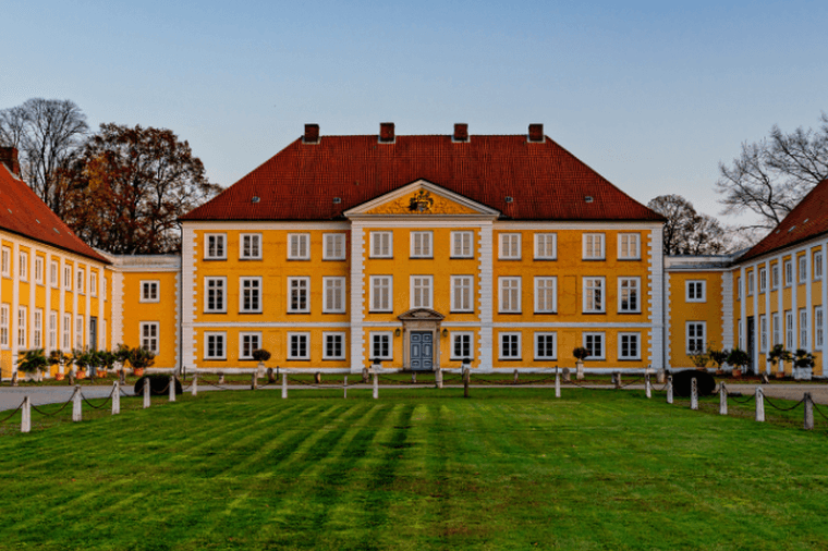 Ein gelbes, historisches Anwesen mit rotem Ziegeldach, einem zentralen Giebel und zwei Flügeln, das hinter einem gepflegten grünen Rasen und kahlen Bäumen liegt